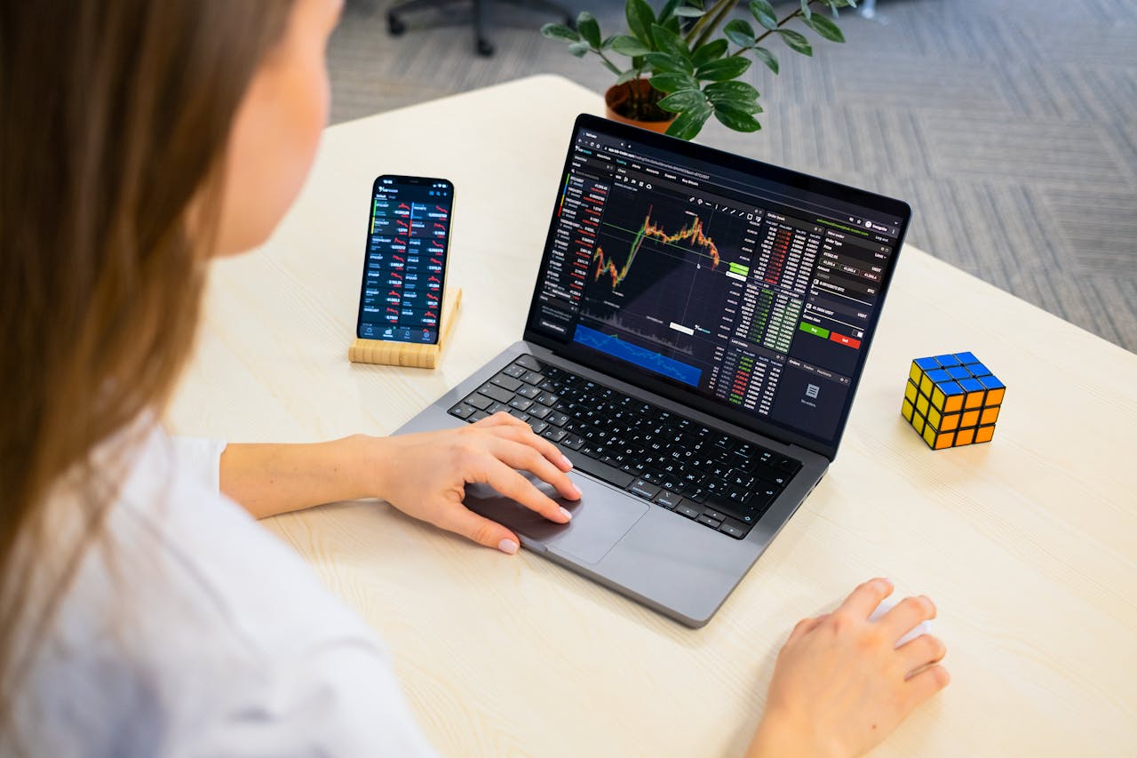 Home A woman using a laptop for stock trading analysis with a smartphone and Rubik's Cube nearby.